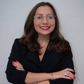 Uma mulher, com idade entre 20 e 25 anos, está sorrindo enquanto olha para a foto. Ela usa uma blusa preta, tem cabelo longo e está de óculos